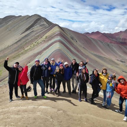 Group of hikers posing with a peace sign and rock-on gestures on the ridge of Rainbow Mountain (Vinicunca), Peru, showing its vibrant red, yellow, and white stripes.