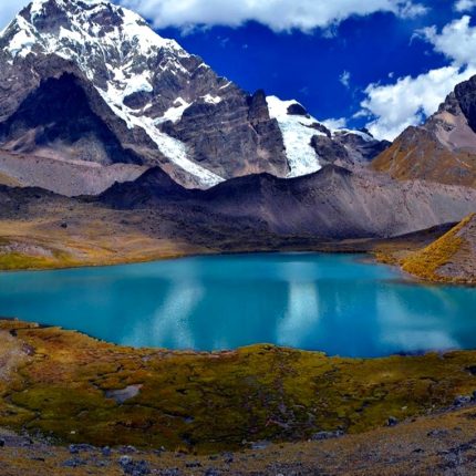 A stunning view of one of the Ausangate Seven Lakes, with a vibrant turquoise-colored lagoon in the foreground and the snow-capped Ausangate mountain peaks towering in the background under a clear blue sky.