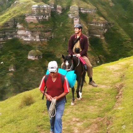 Two people on a Waqrapukara horseback riding tour in Peru, with one man on a horse and another leading the way on a grassy trail. Sheer cliffs and mountainous terrain are in the background.